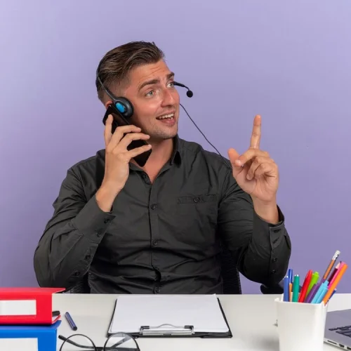 young-surprised-blonde-office-worker-man-headphones-sits-desk-with-office-tools-using-laptop-talks-phone-pointing-up-isolated-violet-background-with-copy-space_141793-65131